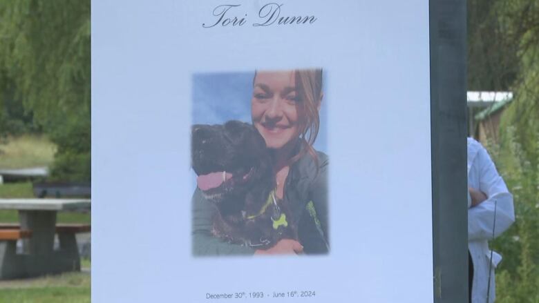 A funerary photo of a white woman smiling with a black dog. Text reads 'Tori Dunn, December 30th, 1993 - June 16th, 2024.'