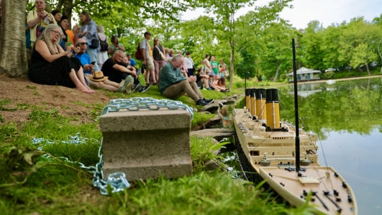 A group of people is seen sitting on a bank with a large Titanic model in the water in the foreground.