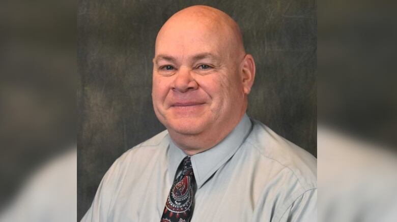 A bald man in a collared shirt and tie faces forward and smiles.