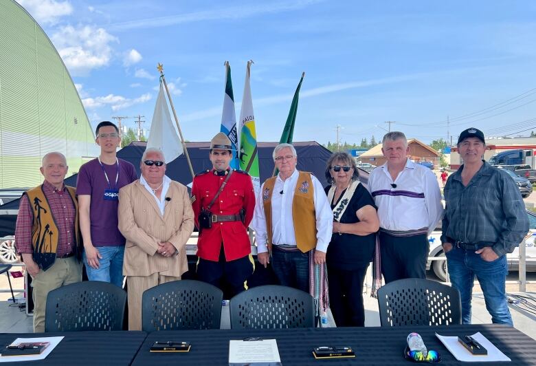 Eight people of varying heights and ages, including one woman, stand side-by-side on an outdoor platform, behind a table where they signed an agreement