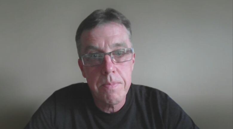 A man wearing a black t shirt and glasses sits in front of a grey wall.