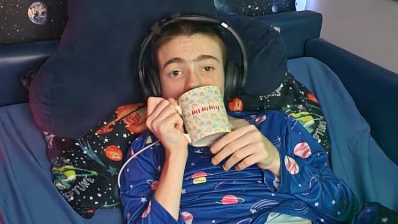 A boy with space pajamas and matching wallpaper lies in bed with a pair of headphones on, sipping from a mug.