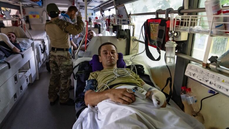 A man in green shirt rests on a stretcher inside a large vehicle while other men work with medial equipment in the background. 