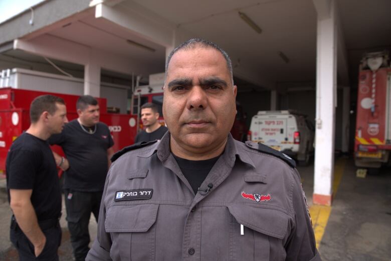 A man with close-cropped greying hair wears a firefighter's uniform and stands in front of an emergency vehicle bay as a group of firefighters speaks behind him. 