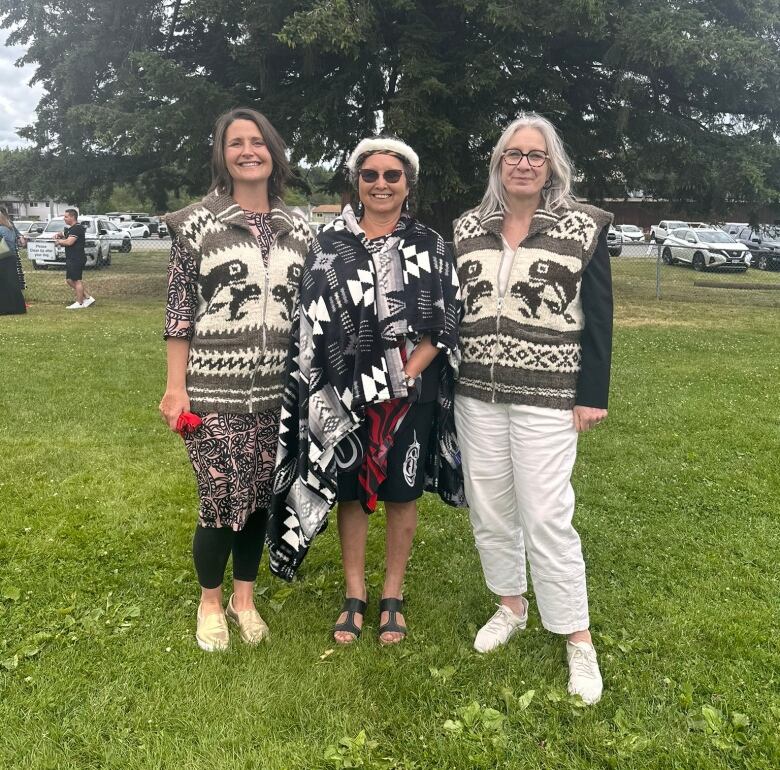 Three women wearing Indigenous regalia smile while outdoors.