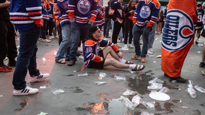 A man sits on the pavement surrounded by empty beer cups and other litter.