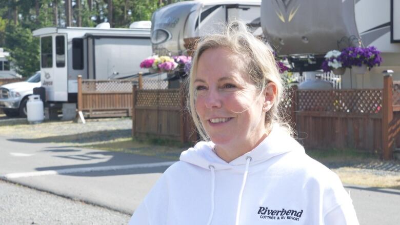 A blonde woman wearing a white hoodie smiles while a row of RVs can be seen behind her.