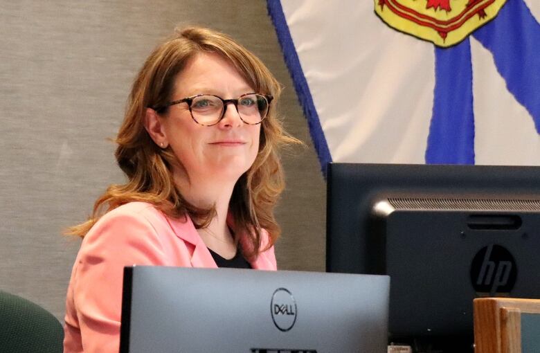 A woman with long reddish brown hair and glasses wearing a pink blazer sits and smiles behind a computer screen with a Nova Scotia flag behind her.