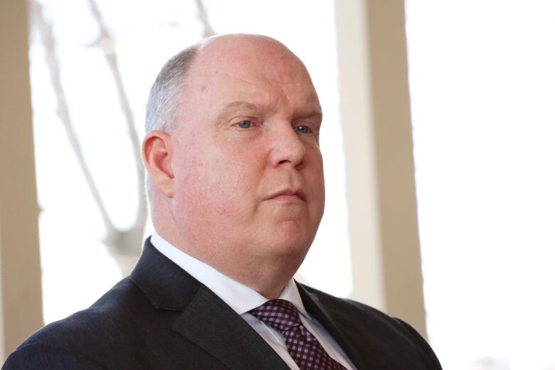 A man in a dark suit and white shirt with a dark red tie stands in front of a window.