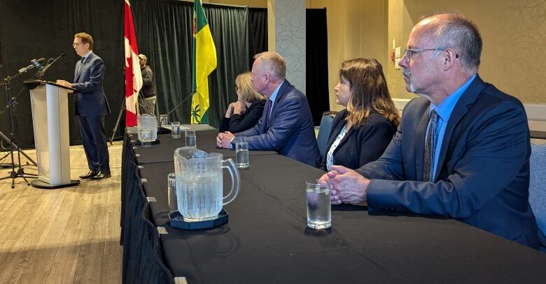 People sit at a table with a man at a podium speaking into a microphone