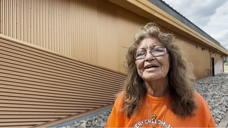 A woman with glasses and brown hair just past her shoulders wearing an orange shirt. Behind her is a wooden building. 