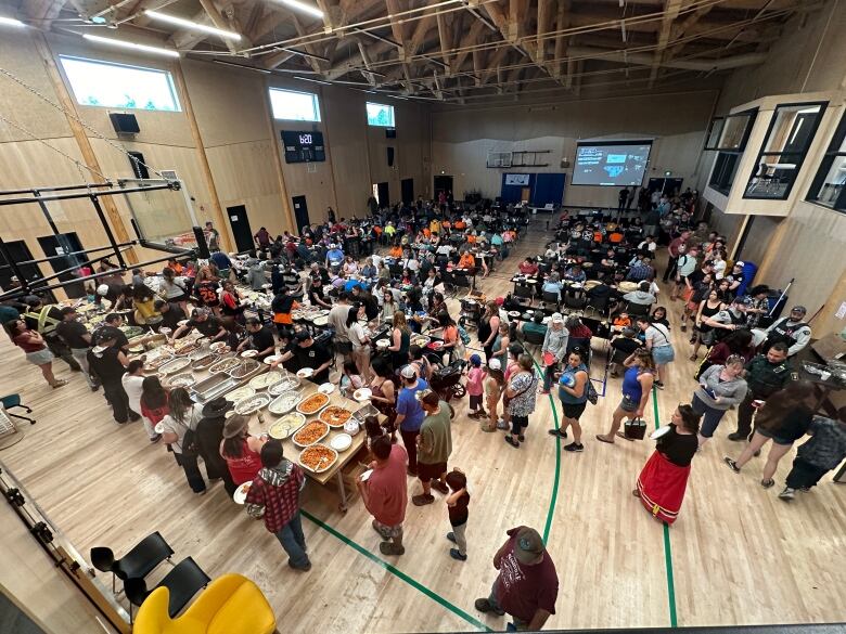 Dozens of people standing in a long line on a wooden gym floor. There's a serving table with several large trays of food. 