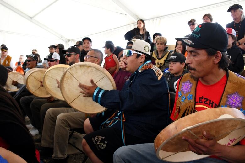 Several men sit in a row playing hand drums. Observers sit in the background. 