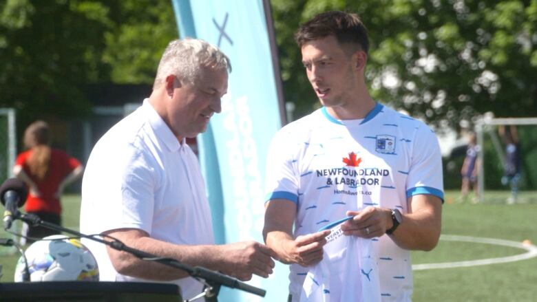 Two men wearing white shirts stand on a soccer field.