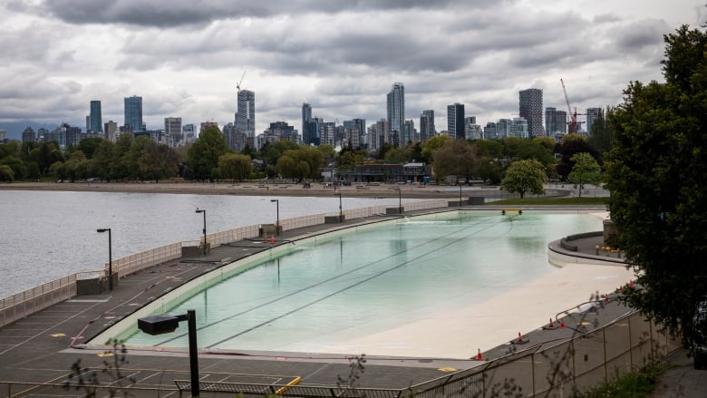 An outdoor swimming pool lies next to the ocean, with a cityscape of downtown office towers and buildings in the background.