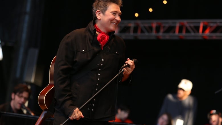 A woman in a black shirt with red scarf smiles on stage. She has a guitar on her back. 