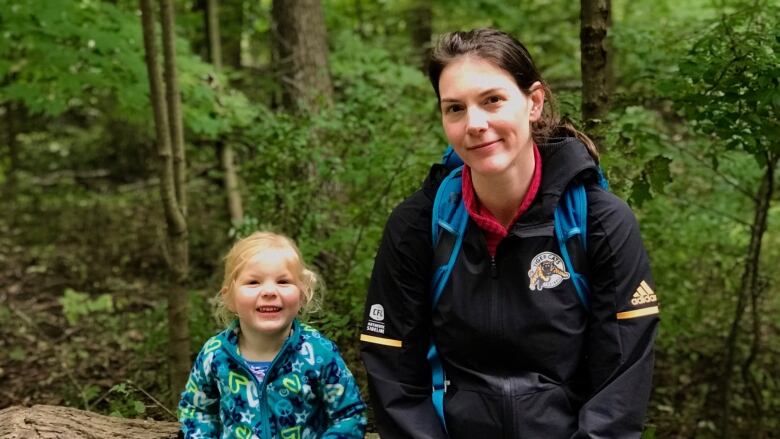 A kid and mom in a Ticats jacket stand in a very green wooded place.