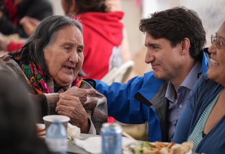 An older Indigenous woman speaks to a white man at a table, with other people visible in the background.