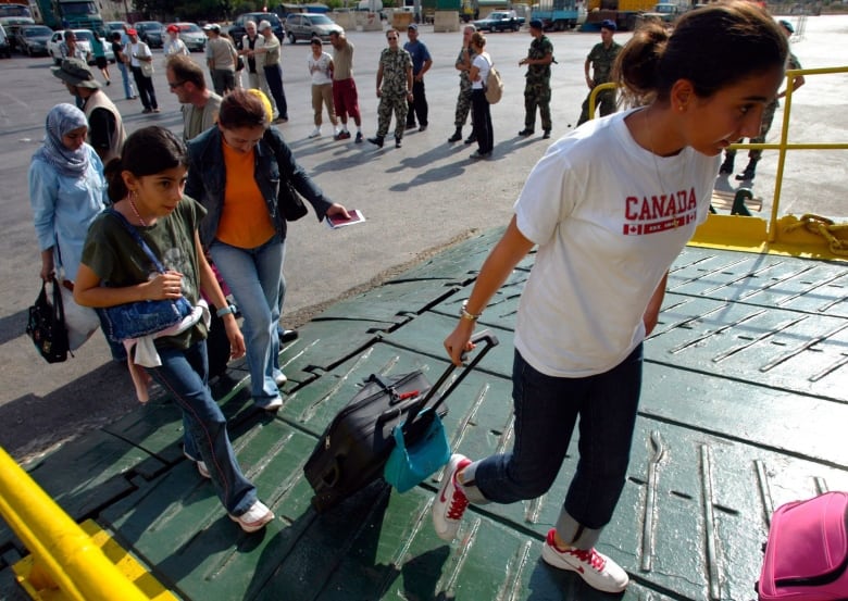 A woman wearing a white Canada t-shirt drags a suitcase up a ramp as she and others board a ferry. 