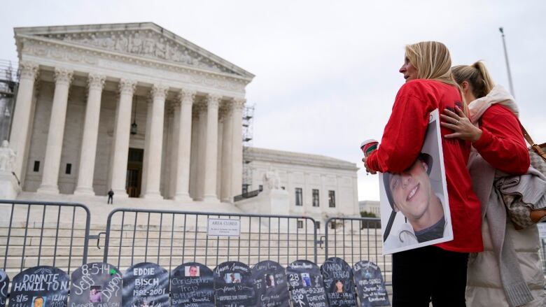 A woman stands in front of an august building, with a picture of someone under her arm. Nearby are makeshift memorials to several people.