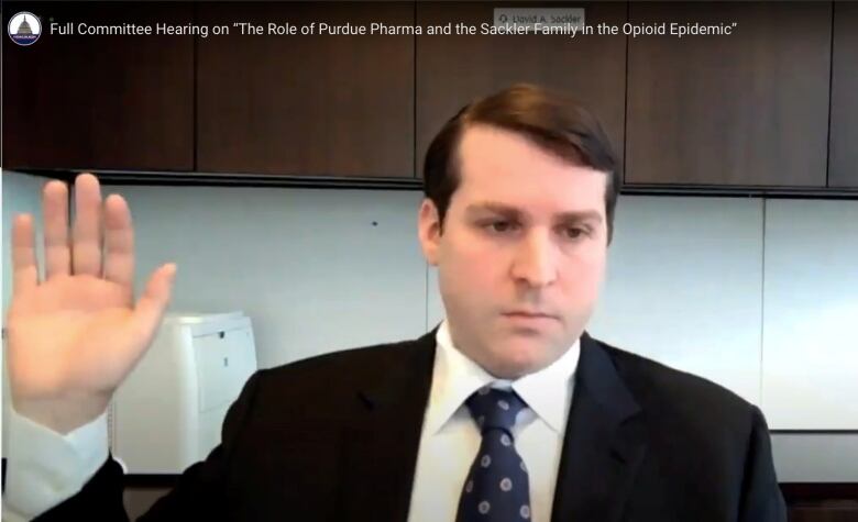 A cleanshaven man in a suit and tie is shown raising his right hand while sitting at a desk in a still taken from video.