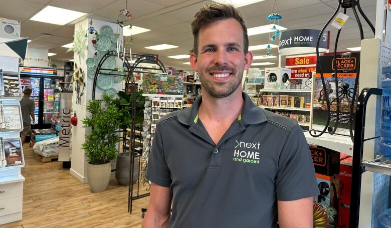 A man who looks to be in his 30s stands in a hardware store with shelves full of gardening supplies and tools. 