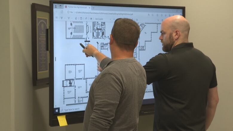 Two men, one bald with a beard wearing a black shirt, the other with short cropped hair and a grey shirt, pointing to floor plans of a school on a monitor.