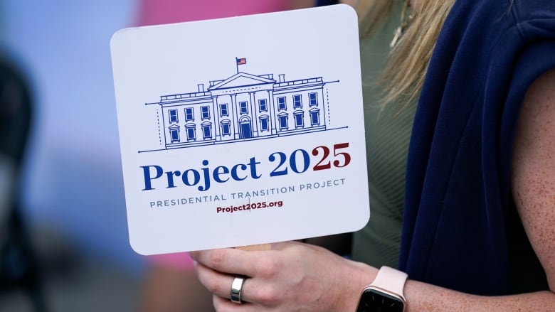 A person, wearing wearing an Apple Watch on her wrist and a gold ring on her finger, holds up a sign with an image of the White House on it and the words 