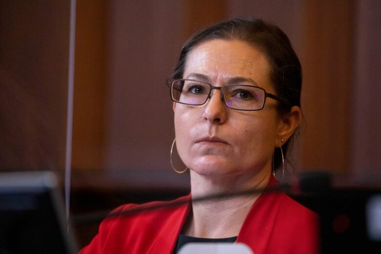 A portrait of a white woman with dark hair pulled back, wearing dark-framed glasses and large gold hoop earrings.