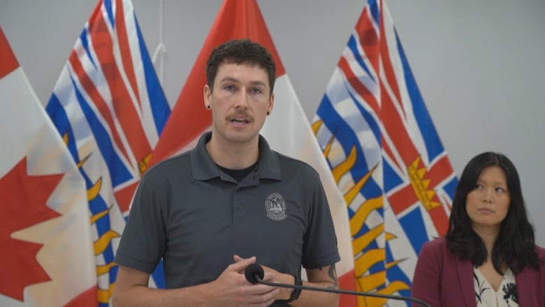 A man speaks at a podium in front of B.C. and Canadian flags while a woman looks on.