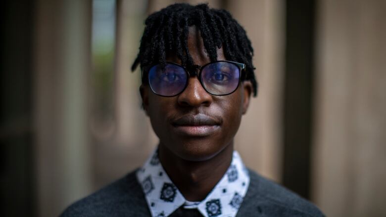 A Black man with dreadlocks and glasses, wearing a collared shirt underneath a sweater, looks at the camera.