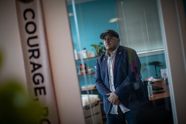 A man in a baseball cap wearing a shirt and jacket stands in a doorway, part of which has the word Courage on it.