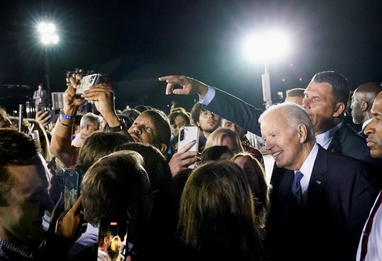 A man is shown smiling while surrounded by dozens of people on the tarmac of an airport.