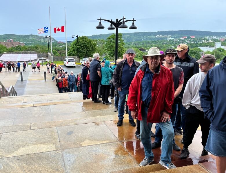 Several dozen people are in a line that stretches down a long series of steps. Flags in the background are at half-mast. 