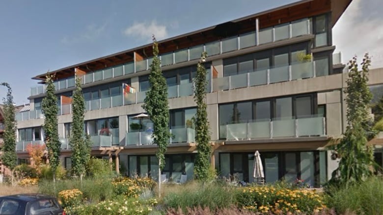 A long, four-storey condo building fronted by trimmed greenery and a sidewalk where two cars are parked.