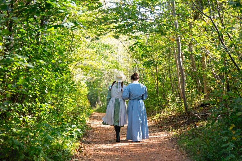 Two women in old-fashioned clothing walking down a treed path. 