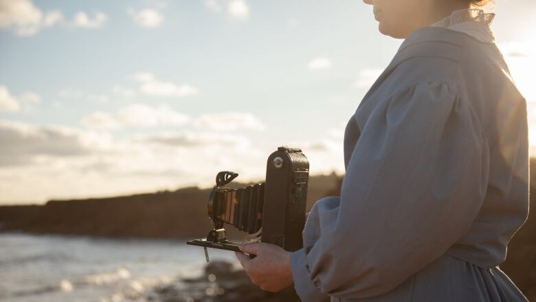 A woman wearing an old-fashioned dress holding an old camera 