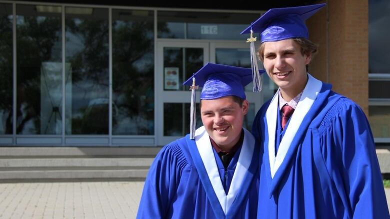 Two young men in blue graduation gowns and mortarboards smile as they stand outside a school.