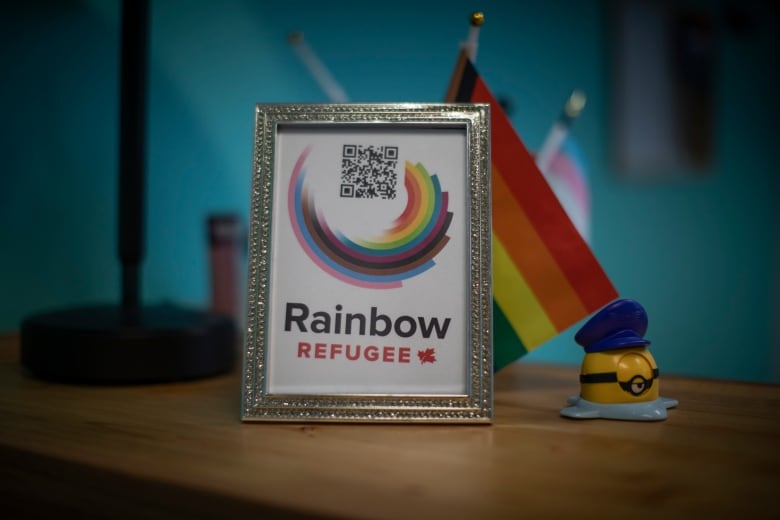 A rainbow flag is seen behind a silver-framed plaque that says Rainbow Refuge next to a small Canadian flag  all of which sits below a curved rainbow semi-circle.