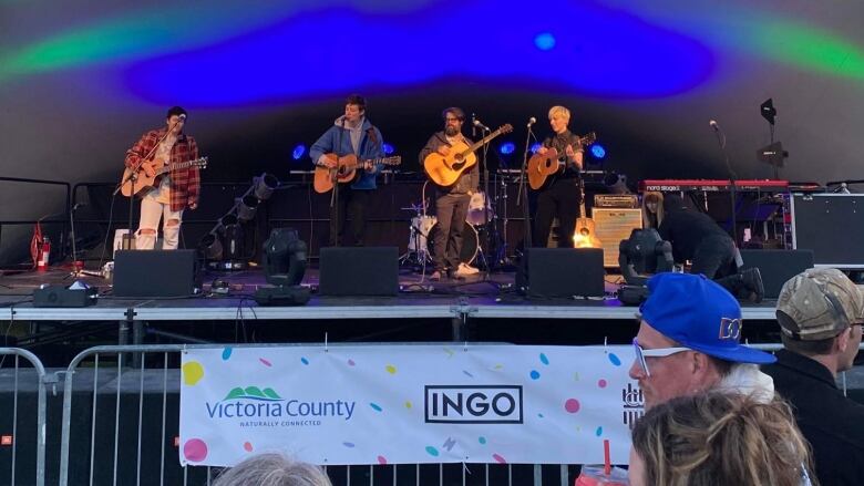 Four musicians perform on stage. In front of them is a sign that reads INGO. Several people can also be seen in the crowd. 