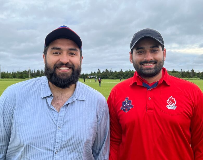 Two men smile. Behind them, people play cricket on a field.