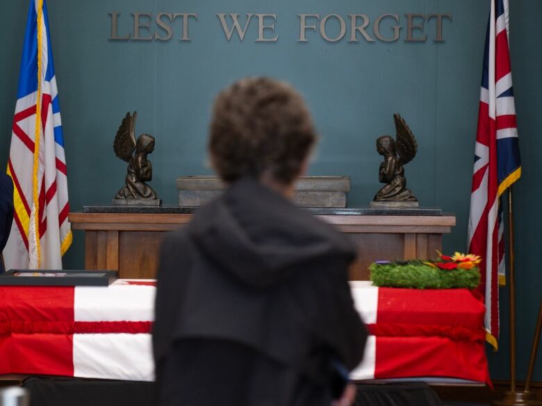 A young person wearing a dark hooded sweatshirt bows their head before a casket draped in the Canadian flag. 