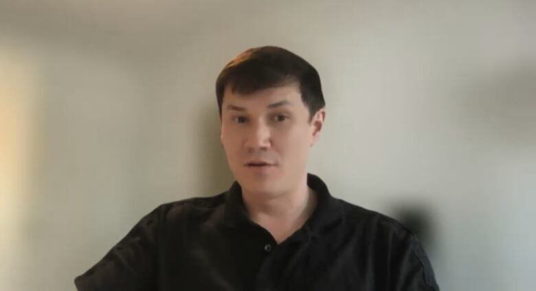 A man in a black polo shirt sits for a portrait in front of a blurred background. 