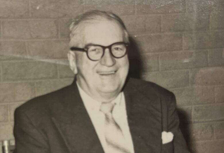 a black and white photo of an older man in a suit wearing glasses and smiling.