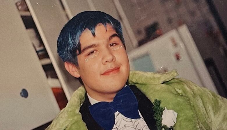 A person in a blue bowtie and green textured jacket, with gemstones under their eyes. 