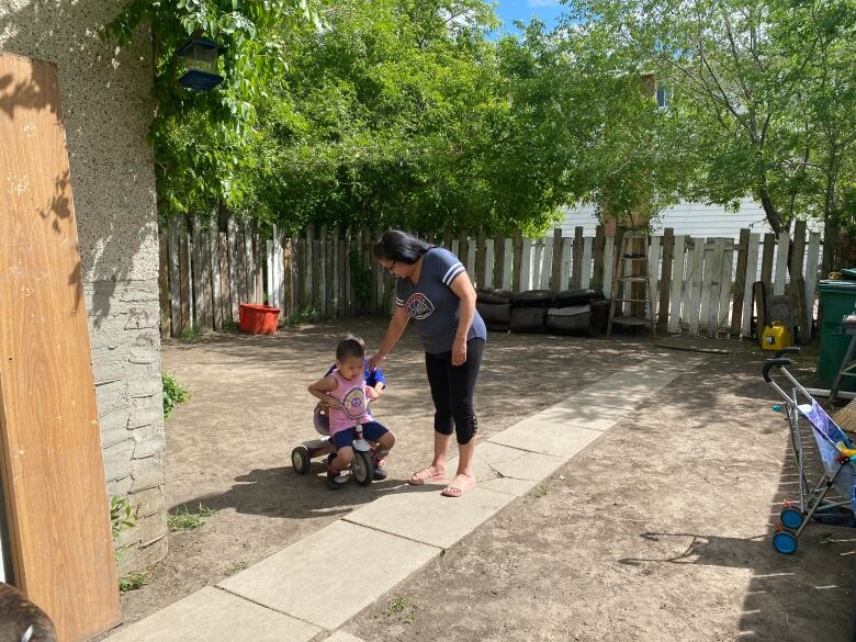 A black-haired woman stands on a concrete path in a backyard, with two children balancing on a tricycle. 