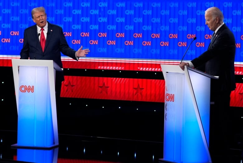 A man in a suit on the left stands behind a podium, that is light in a blue-white gradient, motioning with his left hand toward another man in a suit standing behind a similar podium.