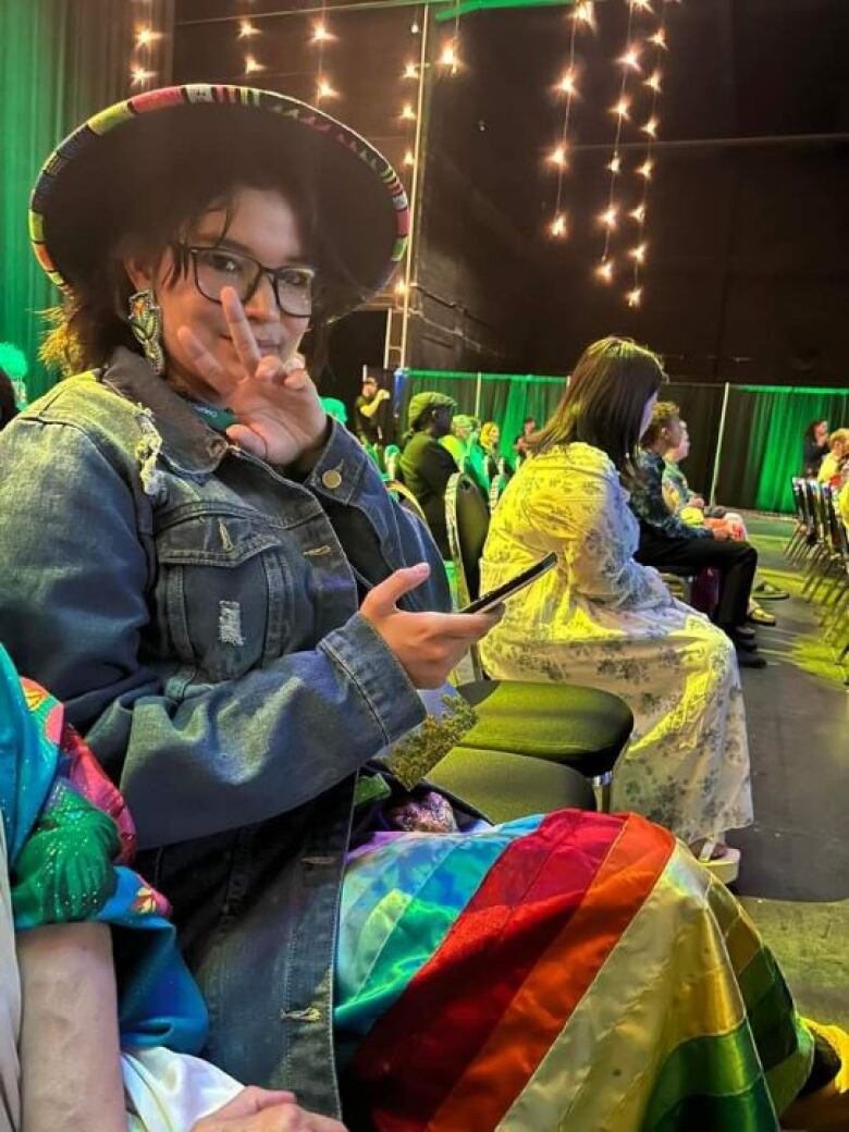 A woman wearing glasses and a colourful ribbon skirt and denim jacket flashes a peace sign, with people on a stage seen behind her.