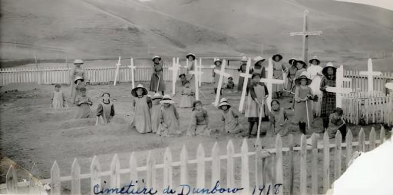 Roughly two dozen children gather in a cemetery, standing tamid wooden crosses marking what look like shallow, small graves of their peers.