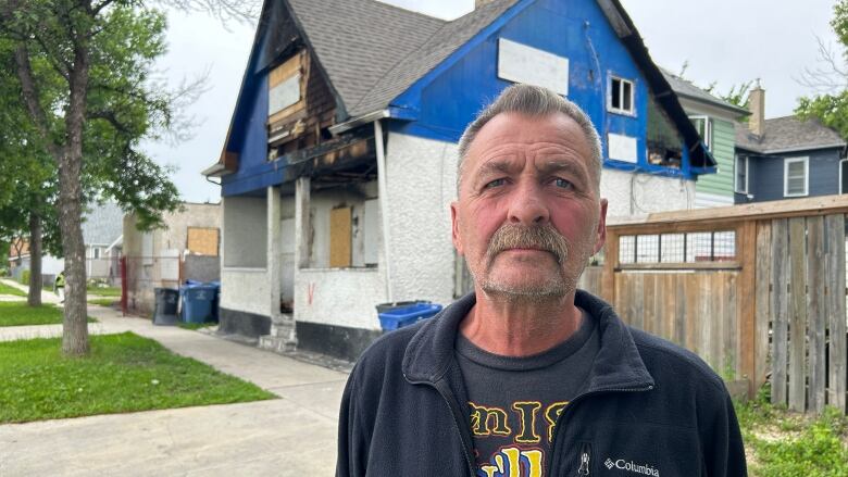 A man wit ha mustache is standing in front of a burned-out house.